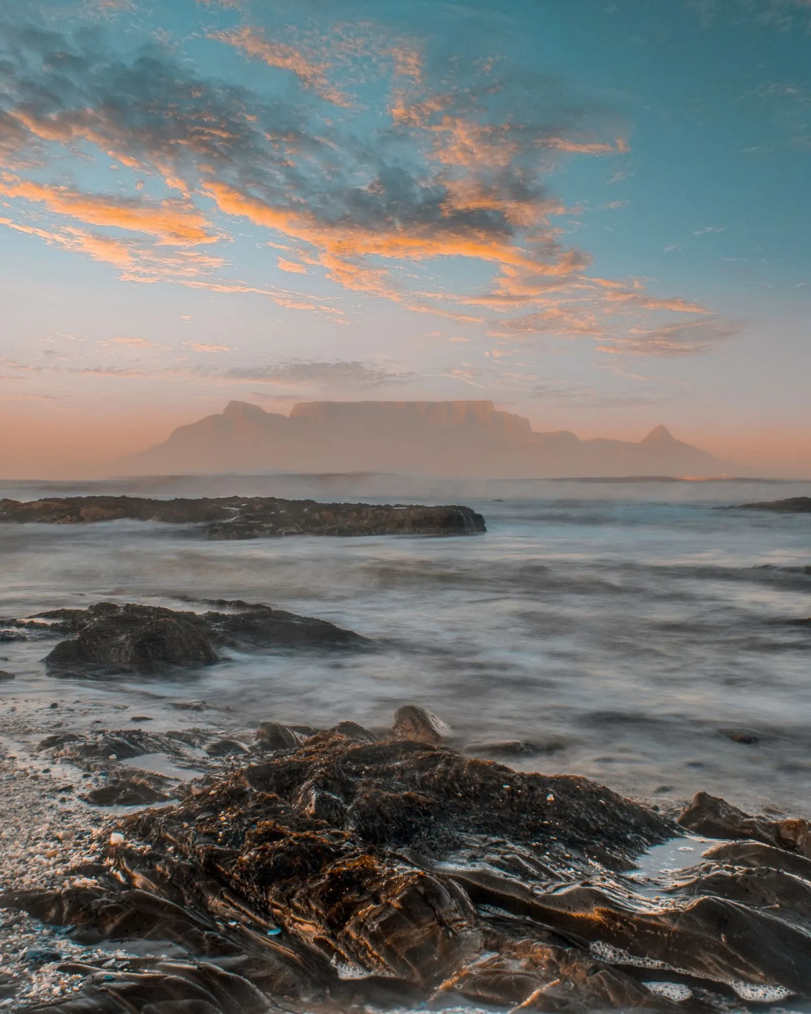 Table Mountain in evening haze beyond surf washing over dark rocks, orange clouds in a teal sky