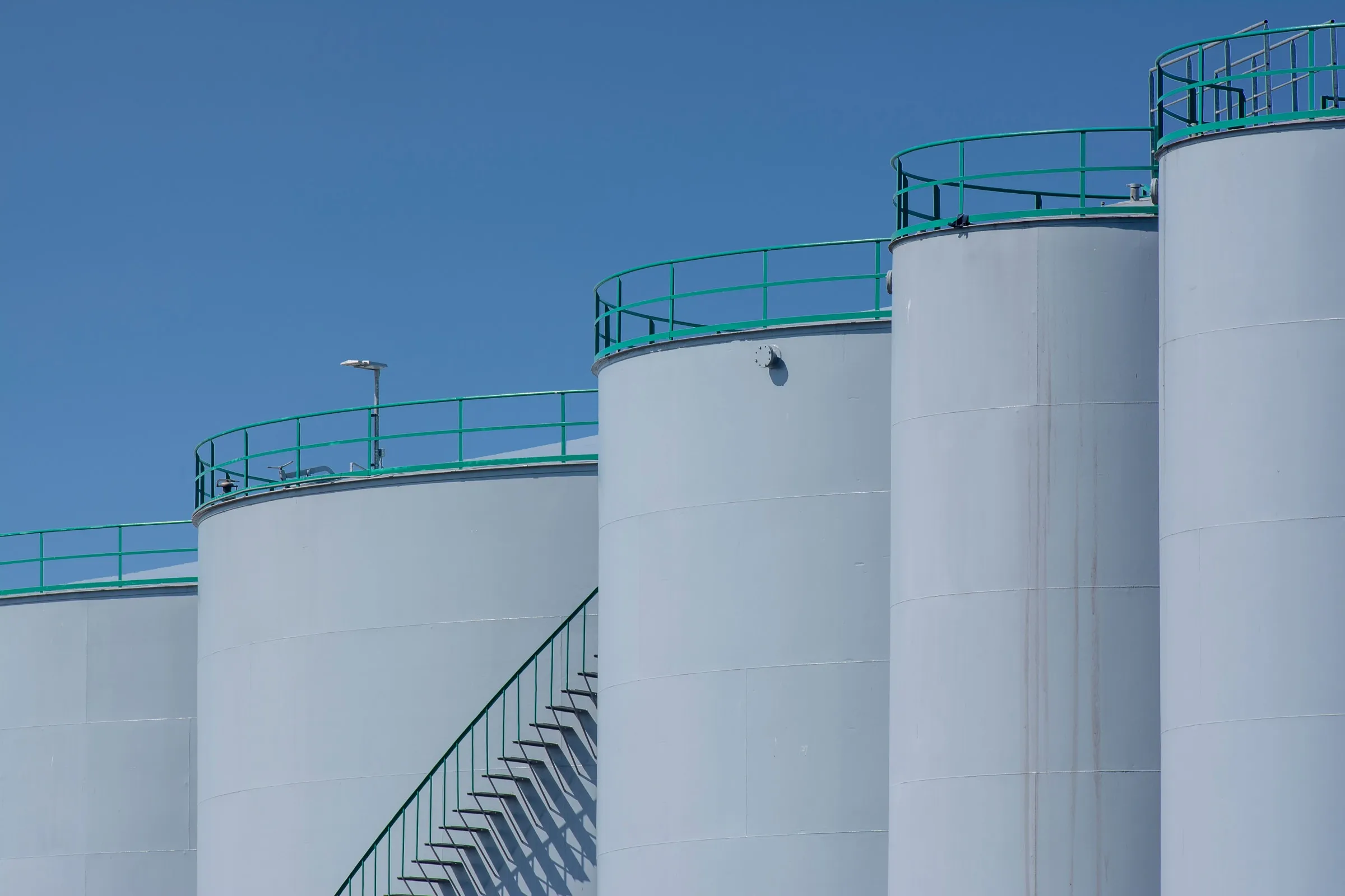 Tall grey storage tanks with teal railings against a blue sky